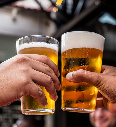 Two hands clinking glasses of beer in a toast, with golden liquid and frothy foam visible. The background is slightly blurred, showing an outdoor or bar setting with people, including a smiling man.