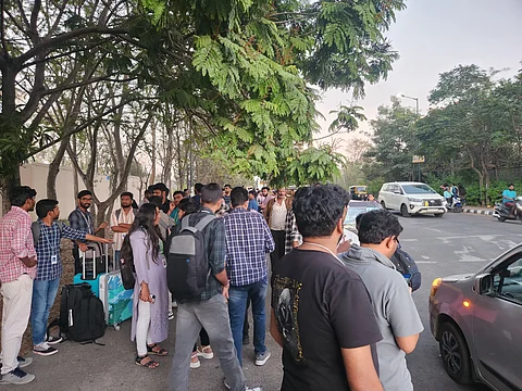 The terminated employees, some carrying backpacks and suitcases, stand on the side of a tree-lined road. Some individuals are engaged in conversation, while others appear to be waiting.