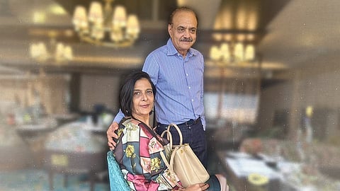An image of Pooja Singh (seated, wearing a multicoloured dress and holding a beige bad) and Shyam Sunder Bhartia (standing next to her, right hand on her shoulder, wearing a blue shirt). The background of the image has been blurred.
