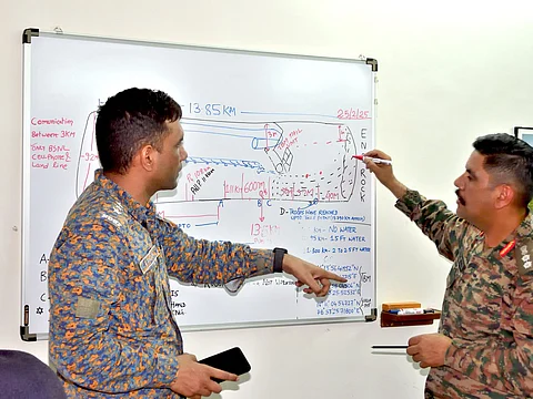 Two military personnel in camouflage uniforms are engaged in a discussion in front of a whiteboard filled with diagrams, notes, and measurements. One officer, holding a smartphone, is gesturing towards the board while the other is marking details with a red marker. The board contains tactical information, distances, and water levels, suggesting a briefing or planning session related to a rescue or military operation. The date "25/2/25" is visible on the board, along with various annotations and coordinates.