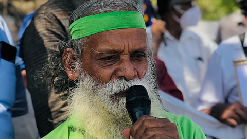 An elderly man, environmentalist Kallur Balan, with a long white beard and a green headband is speaking into a microphone at an outdoor event. He is wearing a bright green polo shirt. Behind him, several people, some wearing masks, are gathered, with banners and flags visible in the background. The setting appears to be a protest or public gathering.