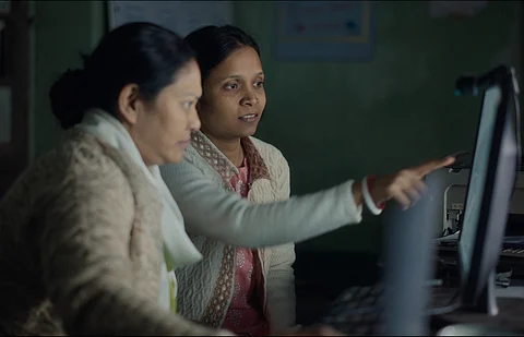 Two women look at a computer screen. we are shown their side profile, and cannot see what's on the screen. One of the women is pointing at the screen. Both are wearing cardigans.