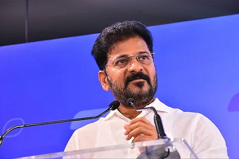 Revanth Reddy with a beard and glasses, wearing a white shirt, is standing behind a podium with two microphones, speaking at an event. The background is blue with a subtle design.