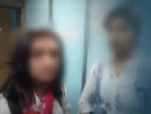 A blurred image of two girls standing indoors, wearing backpacks and casual clothing