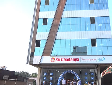 A modern multi-story glass building with a blue and white facade, featuring a diagonal brown panel. The entrance is decorated with blue and white balloons, and several people are gathered near the doorway. The signage above the entrance reads "Sri Chaitanya Academy," along with "Knowledge Hub" and "Infinity Learn." The surrounding area includes older structures and an open sky in the background.