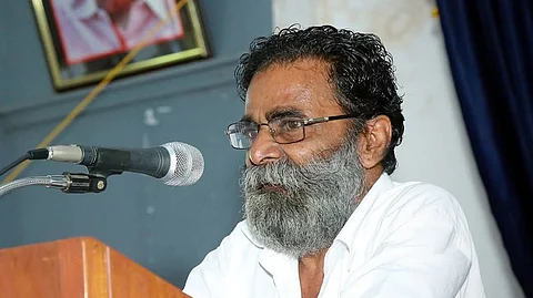 KK Kochu wearing glasses and a white shirt speaks into a microphone at a podium.