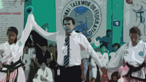 A referee in a white shirt and tie raises the arm of a martial artist in a white dobok with a black belt, signaling victory in a Taekwondo match. Another competitor stands on the other side. The background features a crowd and a large banner with the words 'Taekwondo Federation.'