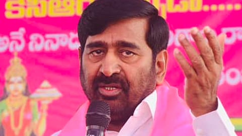 The image shows BRS MLA G Jagadish Reddy wearing a white shirt and a pink scarf with party symbols and text, speaking into a microphone at a public event. He has a serious and expressive facial expression, gesturing with one hand while addressing an audience. The background contains a pink banner with Telugu text and images of other people, likely related to a political gathering.