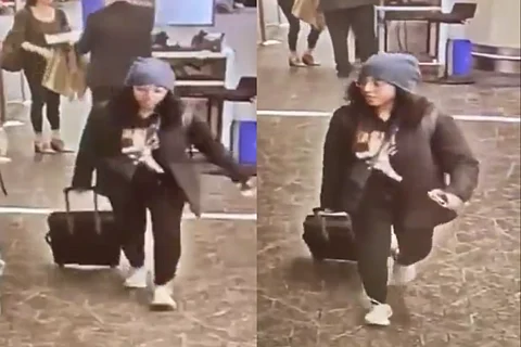 A woman dressed in an all black attire and a blue beanie, walking with a black suitcase.