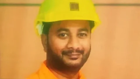 A man wearing an orange safety uniform and a yellow hard hat, smiling slightly at the camera. The background appears to be an indoor setting with a wooden or beige-colored wall.