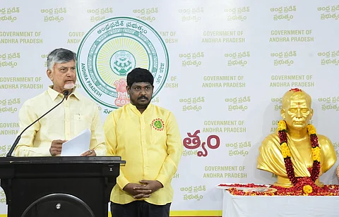 A formal event featuring Andhra Pradesh CM Chandrababu Naidu wearing a yellow shirt standing in front of a backdrop with the emblem of the Government of Andhra Pradesh. Naidu is speaking at a podium with a microphone, holding a document, while another man on the right stands attentively. To their right, a golden bust of Potti Sriramulu is adorned with flower garlands, placed on a decorated table with rose petals. The text on the backdrop and decorations indicates an official government function.