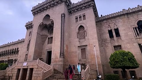 The image shows the historic Osmania University stone building with Indo-Saracenic architectural elements, featuring arched windows, intricate jali (lattice) work, and decorative parapets. A grand staircase leads up to the entrance, where a group of people dressed in traditional Indian attire is walking up. The sky is overcast, and the surrounding area is adorned with greenery.