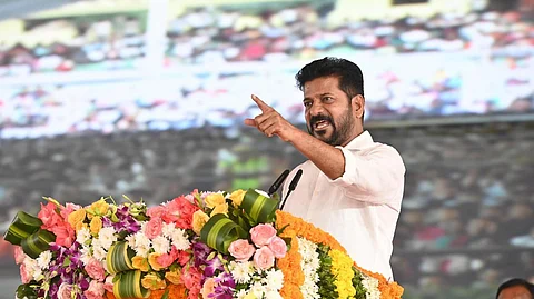 Telangana CM Revanth Reddy in a white shirt is delivering a speech at a public event, standing behind a podium decorated with colorful flowers. He is gesturing with his right hand while speaking, and the background shows a large crowd and a big screen displaying the event.