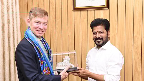 The image shows two men standing in a formal setting. McDonalds CEO Chris Kempczinski, dressed in a suit with a blue and gold shawl, is holding a glass-encased decorative object depicting a Nandi (sacred bull). Telangana CM Revanth Reddy, wearing a white shirt and black pants, is handing over the gift while smiling. The background consists of a wooden panel wall with a framed photo at the top.