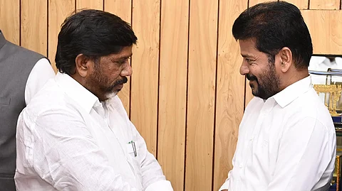 Telangana Finance Minister Bhatti Vikramarka and Chief Minister Revanth Reddy in white shirts are shaking hands in a formal setting. They are looking at each other, with one smiling. The background consists of wooden paneling, and there is a bouquet of flowers in the foreground.