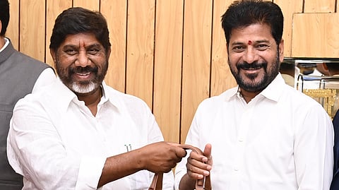 Telangana Finance Minister Mallu Bhatti Vikramarka and Chief Minister Revanth Reddy dressed in white shirts are standing together, smiling at the camera. Bhatti Vikramarka is handing over a brown leather briefcase to Revanth Reddy. The background features wooden paneling and a framed picture of Gandhi. The setting appears formal, possibly an office or meeting room.