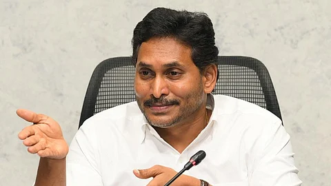 YSRCP chief YS Jagan Mohan Reddy with short black hair, a beard, and medium brown skin is sitting at a desk, wearing a white button-up shirt. He is gesturing with his right hand while speaking into a microphone. He has a confident and approachable expression with a slight smile. A Bosch-branded microphone system is placed in front of him. The background consists of a neutral-colored wall, and he is seated in a black office chair with a mesh backrest.