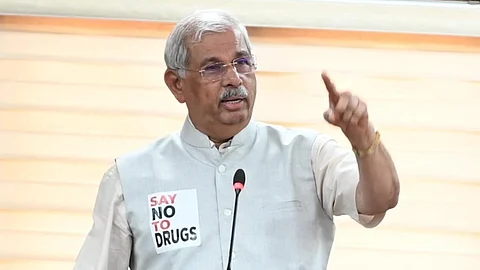 Kerala Governor Rajendra Arlekar is standing at a podium, delivering a speech. He is wearing a beige kurta with a light gray Nehru jacket. A microphone is positioned in front of him, and he is making a gesture with his right hand. On his jacket, there is a badge that reads "SAY NO TO DRUGS." The background features beige blinds, and a security personnel member is partially visible behind him.