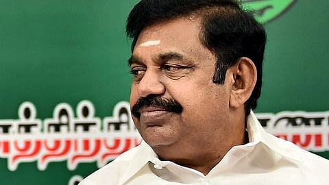 This image shows Edappadi K Palaniswami sitting and looking to his right, with a green backdrop featuring large images of other political figures. The background includes a symbol resembling two green leaves. The text in the background appears to be in Tamil.