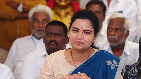 YSRCP leader Vidadala Rajini in a brown blouse and blue saree sitting in front of a bunch of mics addressing the media with her hands clasped together in front of her, with a smile on her face. There are many men in white sitting behind her in the background.