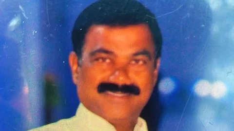 A photograph of a man with short black hair, a thick mustache, and a smile. He is wearing a white collared shirt and is posing against a blurred blue-toned background with hints of light reflections. The image appears to be a printed photo that has been captured digitally.