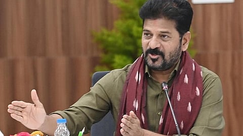 Telangana Chief Minister A Revanth Reddy with a beard and mustache, wearing a green shirt and a maroon scarf with white patterns, is speaking at a meeting. He is gesturing with his right hand while speaking into a microphone. A water bottle and some papers are on the table in front of him. The background features a wooden panel and some greenery.