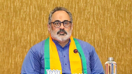 Rajeev Chandrasekhar seated at a table, wearing a blue kurta and a saffron-and-green BJP scarf with lotus symbols, with a microphone and water bottle in front of him.