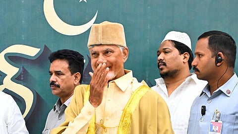 Chandrababu Naidu wearing a traditional golden robe and cap is standing in front of a green backdrop featuring a crescent moon and star. He is surrounded by several men, some wearing traditional white attire and others in uniforms. The man in the golden robe is smiling and appears to be blowing a kiss or offering a respectful gesture. The setting suggests a formal religious event.