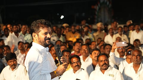 A photograph of Telangana CM Revanth Reddy in a white shirt holding a microphone and addressing a large gathering of people at an outdoor event during the night. The crowd, mostly dressed in white, listens attentively, with some taking photos or recording the moment. Police officers and other attendees are visible in the background, adding to the event's significance. The atmosphere suggests a political gathering.