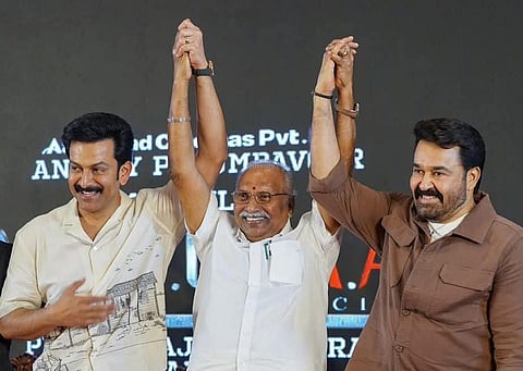 The image shows Prithviraj Sukumaran, Gokulam Gopalan, and Mohanlal standing together on a stage, smiling and celebrating.