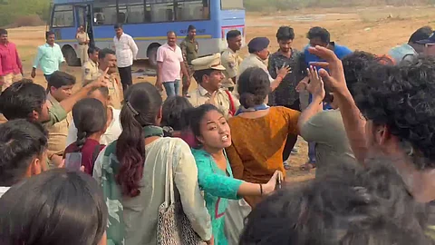 A crowd of people, including police officers and civilians, gather around in an open area near a blue bus. Some individuals appear to be involved in a confrontation or protest, with visible gestures and expressions of distress. A woman in the foreground, wearing a turquoise kurta, is looking toward the camera and extending her arm as if urging someone to stop or follow. The atmosphere seems tense and chaotic.