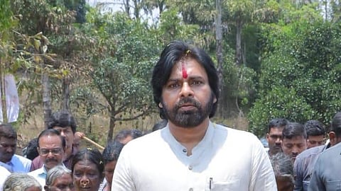 Pawan Kalyan wearing a cream-colored kurta and white pants is walking confidently on a dirt path, surrounded by a group of people. He has a red tilak on his forehead, a small black microphone clipped to his chest, and is wearing sandals. The background is filled with trees and greenery, suggesting a rural or village setting.