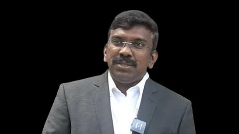 Reny Wilfred wearing a dark grey suit and white shirt, with a microphone clipped to his lapel, standing against a plain white background.