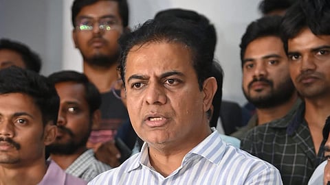 BRS leader KTR in a striped shirt speaks passionately at a press conference, surrounded by a group of attentive people, with multiple microphones from various news outlets positioned in front of him.