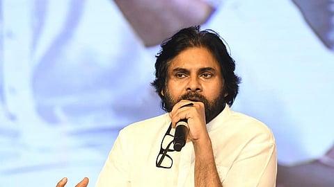 Andhra Pradesh Deputy CM Pawan Kalyan with long hair and a beard, dressed in a white shirt, is speaking into a microphone at a clear acrylic podium. He is holding a pair of glasses in one hand and gesturing with the other. The background features a large, blurred screen showing an image of him.