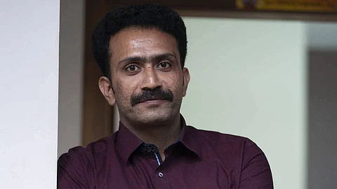 Actor Shine Tom Chacko standing with arms crossed, wearing a maroon shirt and watch, leaning against a wall.