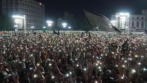 A massive nighttime protest with thousands of people holding up their lit mobile phones, creating a sea of lights. The crowd is densely packed, and some participants wave flags and hold signs. Large stage lights illuminate the area, and prominent buildings are visible in the background.