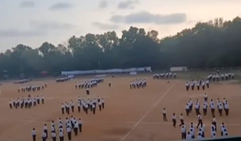 A wide view of an RSS training camp taking place on the Mar Ivanios College ground, showing multiple groups of participants in uniform performing drills in formation under an evening sky, with trees and temporary shelters in the background.