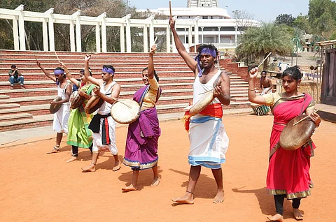 Men and women are playing the parai, a type of percussion instrument, in an amphitheatre. One arm of the men and women is raised.