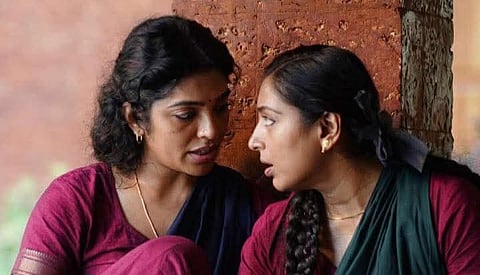 Actors Rima Kallingal and Padmapriya in an intense and emotional conversation, dressed in traditional attire, seated closely against a rustic red brick wall.