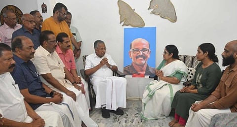 Kerala Chief Minister Pinarayi Vijayan, dressed in white, sits with folded hands, offering condolences to Ramachandran’s family. Seated opposite him is Ramachandran’s wife in a white and green saree, their daughter next to her, and other family members. A portrait of Ramachandran, smiling, is placed between them.