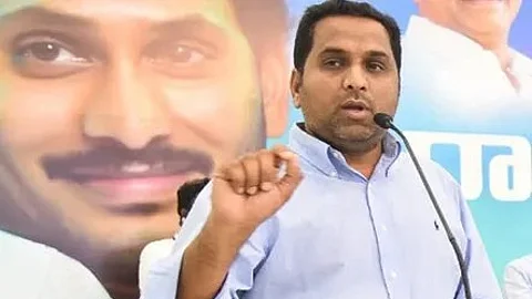 A man in a light blue shirt is speaking into a microphone at a podium. Behind him is a large poster featuring a close-up of YS Jagan's face smiling. The background suggests a political event or press conference.