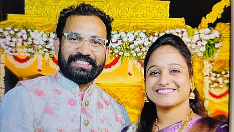 A smiling couple dressed in traditional Indian attire is standing in front of a beautifully decorated golden backdrop adorned with white and red flowers. The man is wearing a floral embroidered vest over a white kurta, and the woman is in a purple and gold silk saree with traditional jewelry.