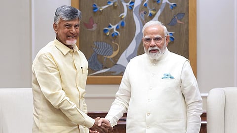 Chandrababu Naidu (left) in a light yellow shirt and trousers, and Narendra Modi (right) in a white traditional outfit, stand indoors shaking hands and facing the camera. A decorative panel with birds and a tree is visible behind them.