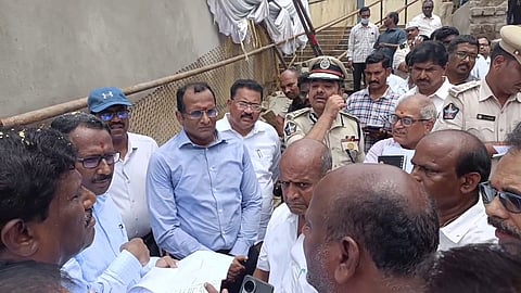 A group of officials and police personnel are gathered in a serious discussion at a construction or inspection site. One officer in uniform appears to be explaining something while holding documents. Other men, including civil officials in formal shirts and some in plain clothes, are attentively listening. The background shows a temporary fence and stairs, suggesting the location is under development or investigation.
