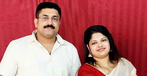 A couple smiling at the camera. Suraj in a white shirt and has glasses. Bincy in a white saree and red blouse. The picture was taken against a red back drop