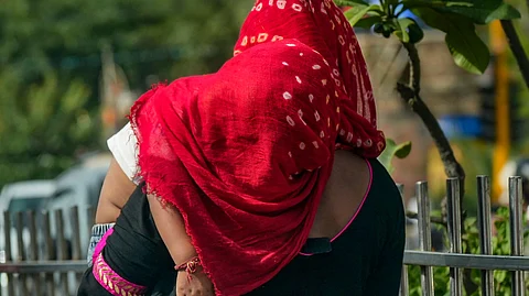 A woman wearing a red dupatta and black traditional attire carries a child in her arms on a sunny day, walking along a metal fence lined with greenery. The child is dressed in white and appears to be resting on her shoulder.