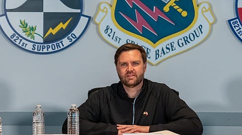 A group of people sit at a conference table in a military meeting room. In the center is U.S. Senator J.D. Vance wearing a black jacket. To his left is a woman in a tan sweater, and to his right is a woman in military uniform. Behind them are five military insignias, including those of the 821st Space Base Group, 821st Support Squadron, and 821st Security Forces Squadron. Water bottles and name placards are on the table in front of them.