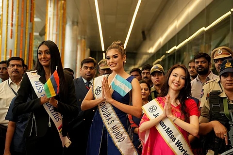 Three Miss World contestants—Miss Mauritius, Miss Honduras Mundo, and Japan Miss World—stand smiling and greeting with folded hands at an airport. They are surrounded by security personnel and others. Each contestant wears a sash indicating their country and holds their national flag.