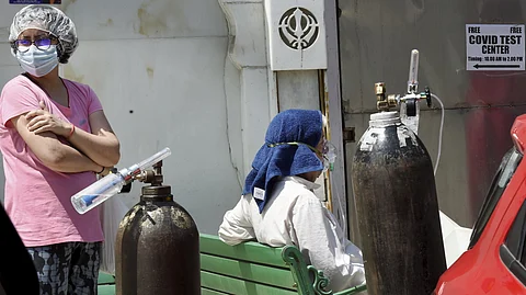 A woman wearing a pink shirt, mask, and hair cover stands with folded arms next to an elderly person sitting on a bench receiving oxygen support. Large oxygen cylinders are placed beside them. The elderly person has a towel draped over their head and is wearing an oxygen mask. A sign on the wall reads "Free COVID Test Center" with operating hours, indicating the setting is during the COVID-19 pandemic.